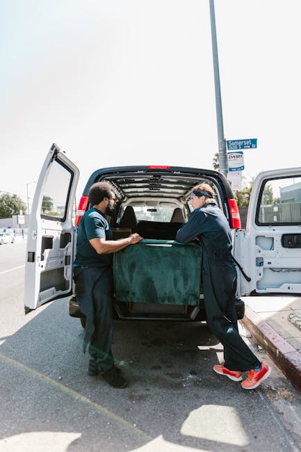 Close-up of a person wearing a dark green t-shirt and blue pants, securing a cardboard box during a home relocation. The individual is applying a caution label on the box that reads 'CAUTION THIS SIDE
