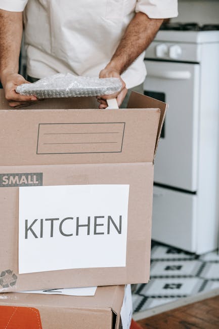 A person wearing a white shirt is seen loading a cardboard box labeled 'KITCHEN' onto a moving vehicle or trolley as part of a home relocation process. The individual is holding a small, protected fragile item wrapped in bubble wrap, preparing it for transport. The scene is set inside a kitchen, with a white stove and patterned flooring visible in the background. The moving boxes are stacked on top of each other, with the labeled box indicating contents related to kitchenware or fragile items that require careful packing and handling during furniture transport or long-distance removals. The image exemplifies the packing and loading process involved in professional removals, emphasizing attention to fragile item safety, and the use of appropriate packing materials such as cardboard boxes and bubble wrap, which are essential for secure moving logistics executed by companies like Man and Van Lee.
