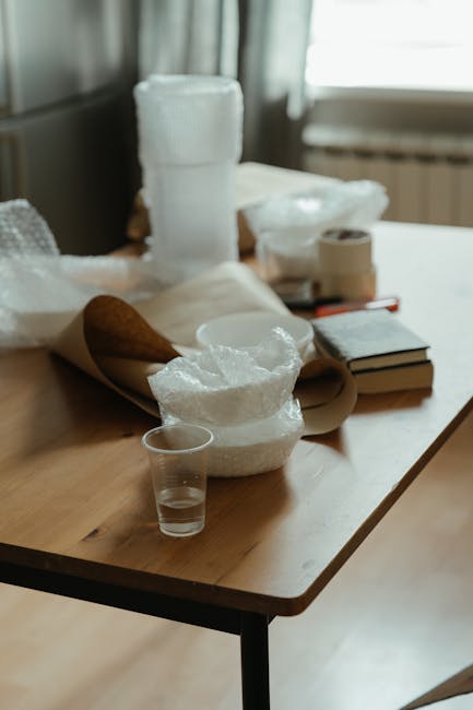 A wooden table indoors holding various packing materials and fragile items for a home relocation, with a stack of white bubble wrap and packing paper at the forefront. Behind these, there are several tightly rolled large sheets of bubble wrap, a small roll of packing tape, and some cardboard boxes. A clear plastic cup is placed near the packing supplies, and the table surface is partly covered with unwrapped fragile objects, possibly dishes or glassware. In the background, windows let natural light into the room, illuminating the scene, while a radiator is visible beneath the window. The setting reflects the process of preparing delicate items carefully for transportation as part of a furniture transport or interior moving task, which a professional removals service like Man and Van Lee might assist with during a long-distance move, especially when packing for fragile items as highlighted in the checklist.