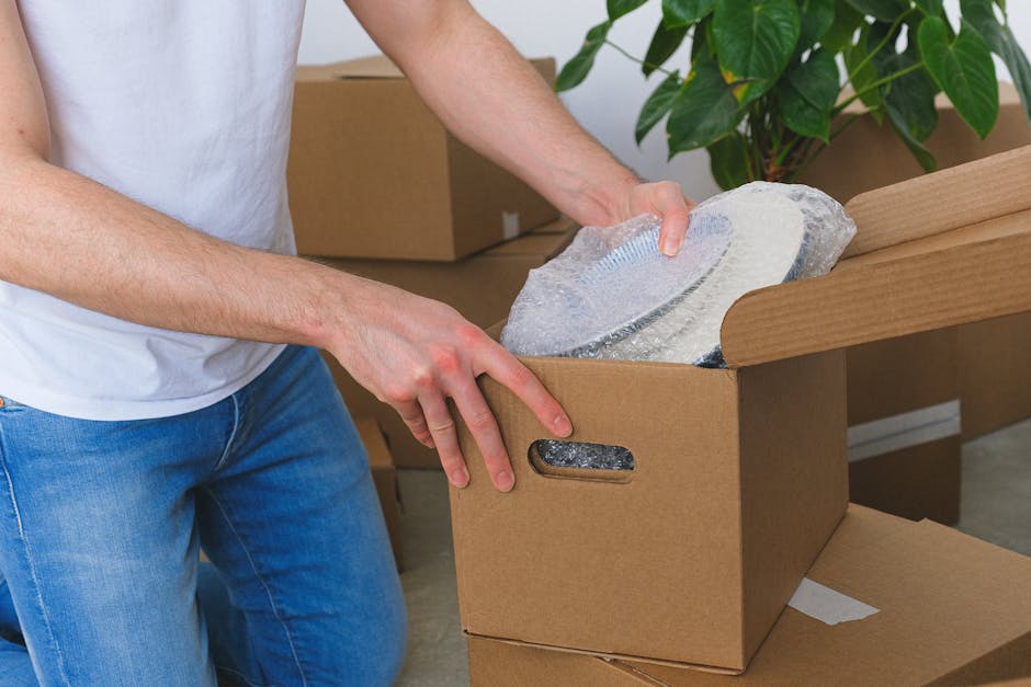 A person wearing a white shirt is seen loading a cardboard box labeled 'KITCHEN' onto a moving vehicle or trolley as part of a home relocation process. The individual is holding a small, protected fragile item wrapped in bubble wrap, preparing it for transport. The scene is set inside a kitchen, with a white stove and patterned flooring visible in the background. The moving boxes are stacked on top of each other, with the labeled box indicating contents related to kitchenware or fragile items that require careful packing and handling during furniture transport or long-distance removals. The image exemplifies the packing and loading process involved in professional removals, emphasizing attention to fragile item safety, and the use of appropriate packing materials such as cardboard boxes and bubble wrap, which are essential for secure moving logistics executed by companies like Man and Van Lee.
