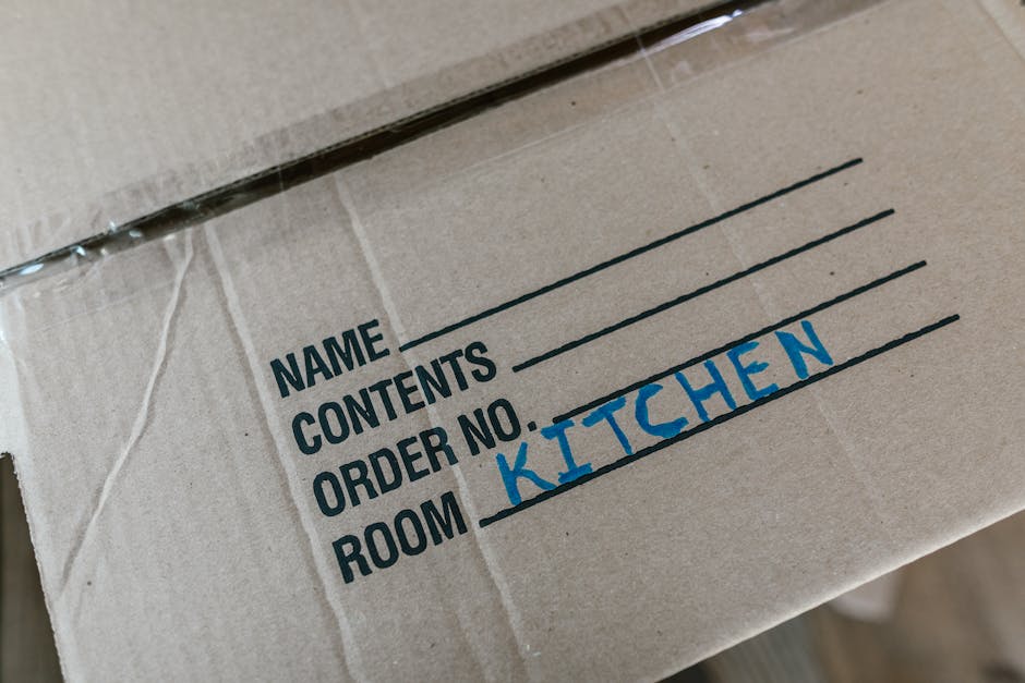A close-up view of a brown cardboard moving box with printed labels indicating spaces for 'Name', 'Contents', 'Order No.', and 'Room', which is handwritten as 'Kitchen' in blue marker. The box appears