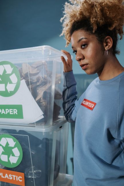 A flat-lay photograph shows a white sheet of paper with a prominent green recycling symbol printed on it, placed on a light wooden surface. Surrounding the paper are various household items related to