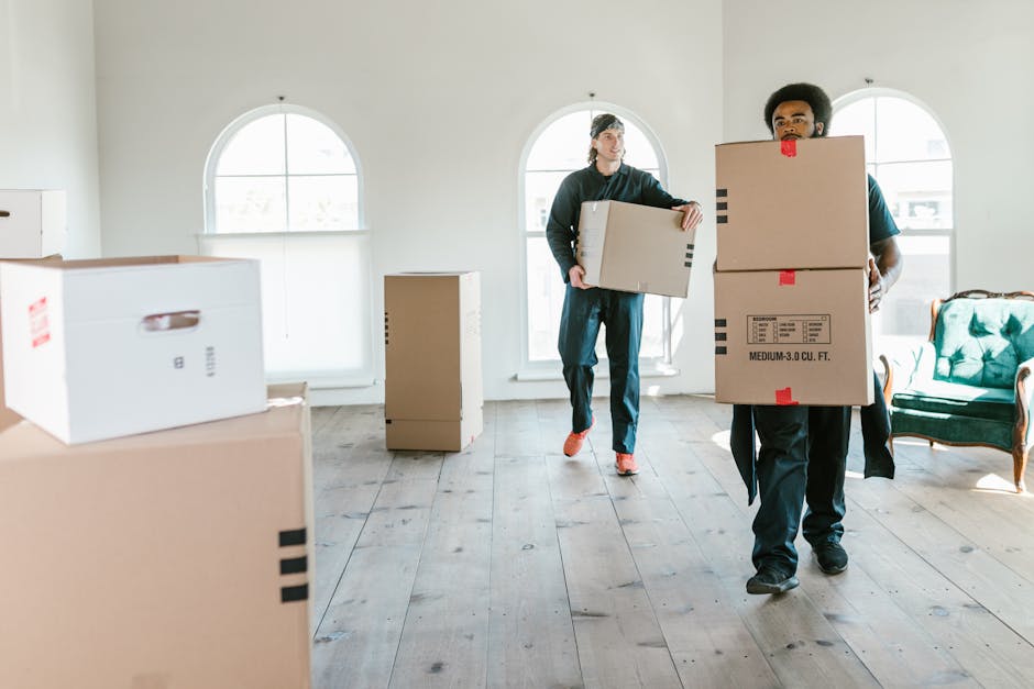 A person wearing a dark green shirt and darker trousers is holding a large cardboard box marked with a red and white caution sticker that reads 'CAUTION THIS SIDE UP' with an upward-pointing arrow. Th